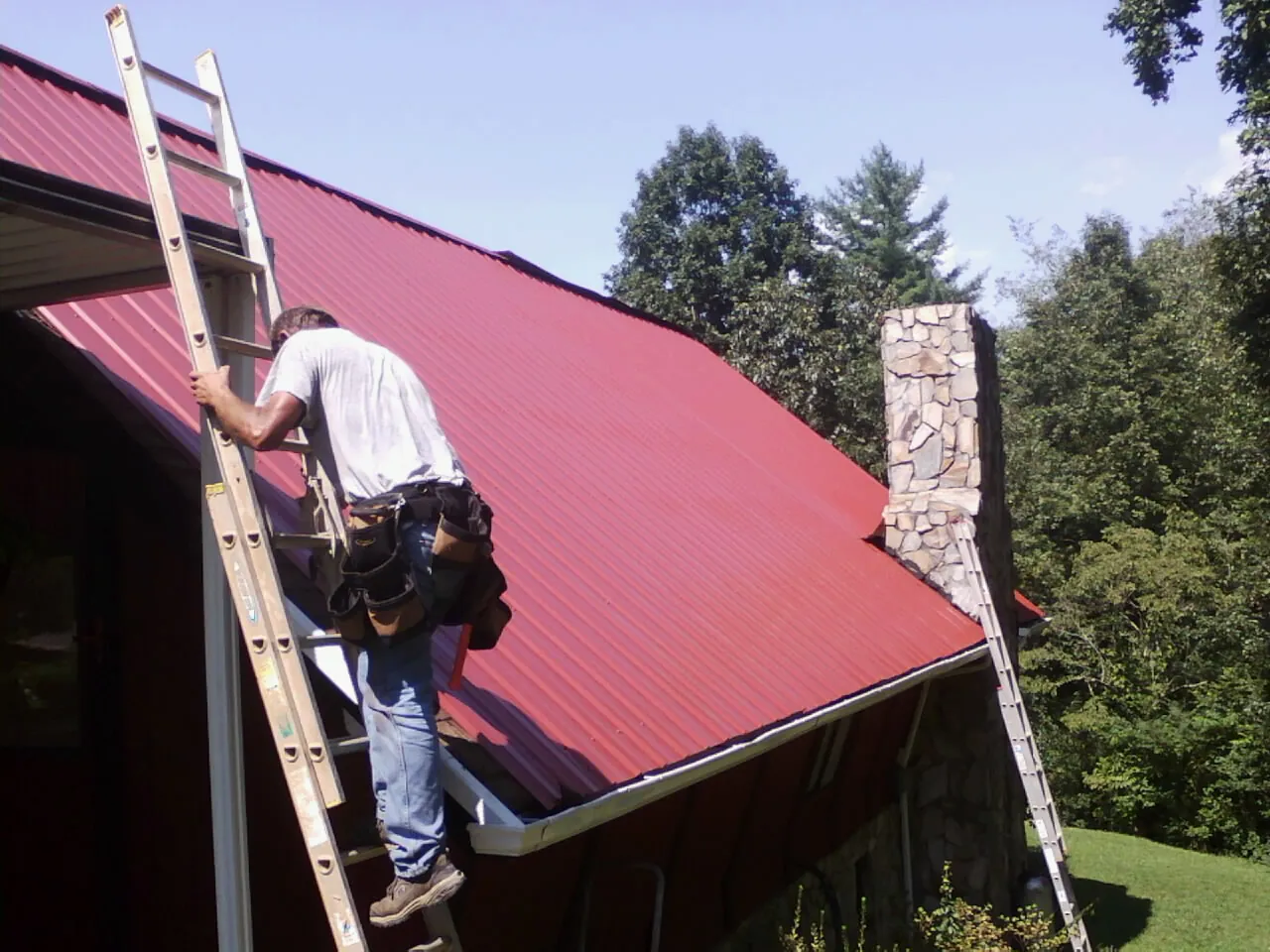 Roofer on a ladder beside a red metal roof and stone chimney — Soffit & Fascia Repair by Roofing Brothers in Gibsonburg